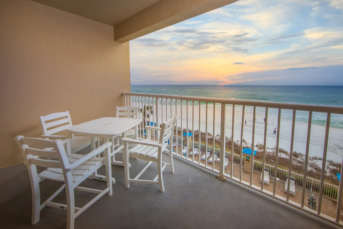 The hero image showcases a private balcony furnished with a white patio set, overlooking the pristine beach shoreline of Panama City Beach. The sun is setting over the Gulf, casting a warm, golden glow across the ocean and creating a tranquil, inviting atmosphere. The scene highlights the seamless connection between the villa and the beach, inviting relaxation and memorable moments right outside the door.