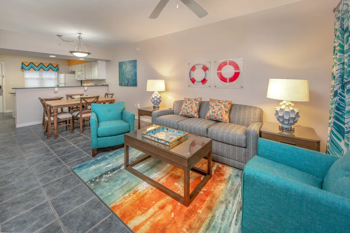 A bright, open-plan living and dining area featuring a striped gray sofa with colorful pillows, two teal armchairs, and a cheerful, multicolored rug. The dining table, set for six with wooden chairs, sits near the kitchen area, making it a natural gathering spot for meals, snacks, and conversation.