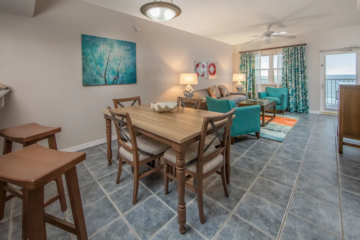 A welcoming villa living and dining space, featuring turquoise armchairs and sofa, a wooden dining table, colorful wall art, and large windows that offer direct views of the ocean. The setting is bright and cheerful, with coastal décor and an open, airy atmosphere.