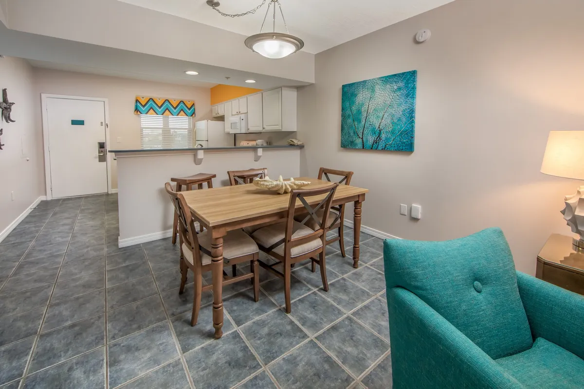 A cozy dining and kitchen area with a wooden table, turquoise armchair, vibrant chevron backsplash, and wall art. The space is set up for socializing, meal preparation, and family gatherings, exuding warmth and personality.