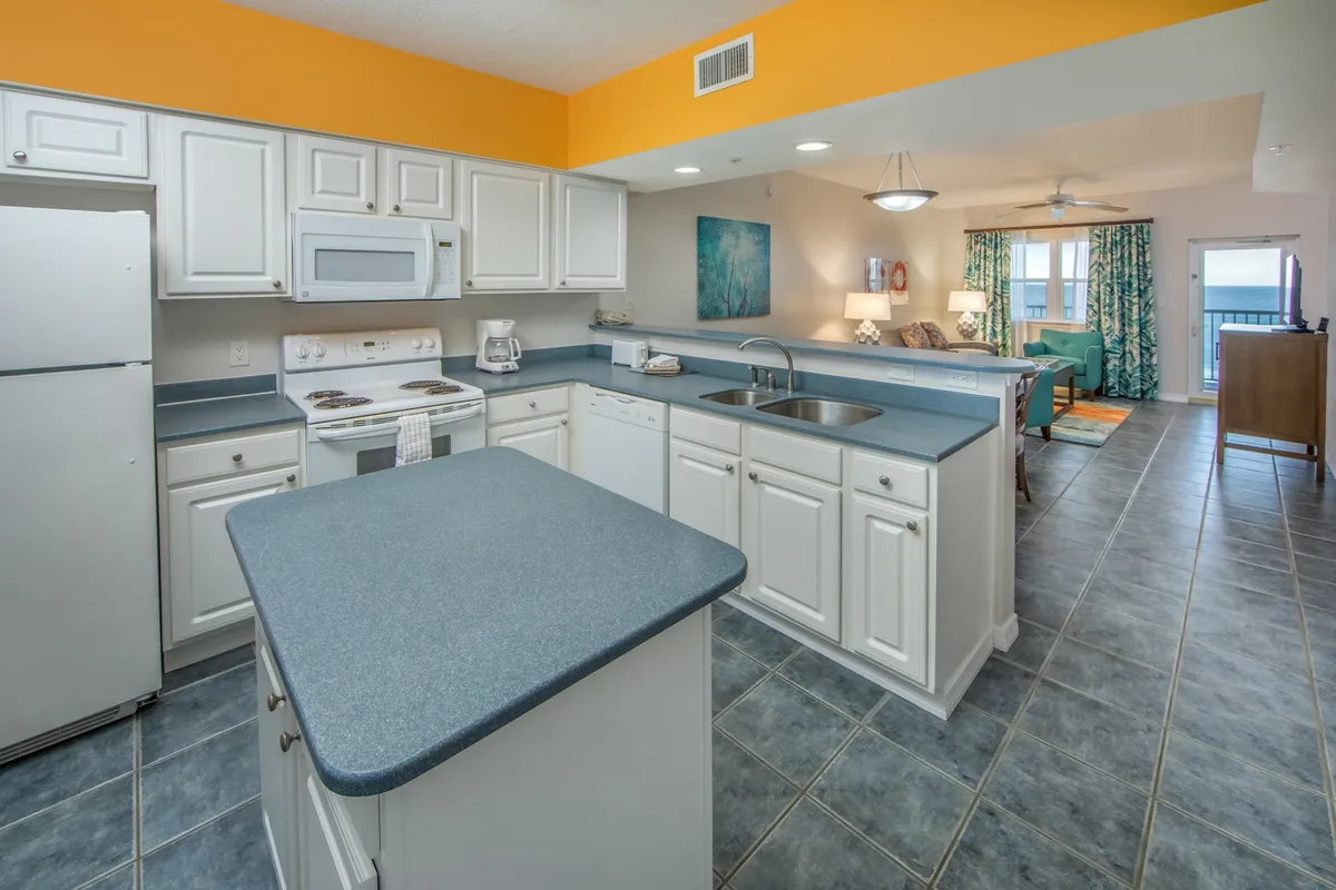 This image features a bright, open-plan living area with a kitchen island, white cabinetry, and colorful furniture. The living space seamlessly connects to a balcony through glass doors, revealing a stunning beach and ocean view. The scene feels relaxed and cheerful, evoking the comforts of a home-away-from-home with easy access to outdoor beauty.