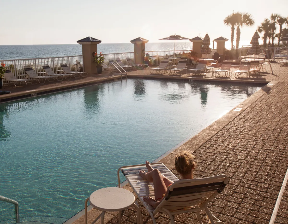 A sunlit outdoor swimming pool overlooks the ocean, with a person lounging on a pool chair under a beach umbrella. The pool area is lined with more chairs and umbrellas, and the ocean horizon glows with the colors of sunset, creating a relaxing, family-friendly atmosphere.