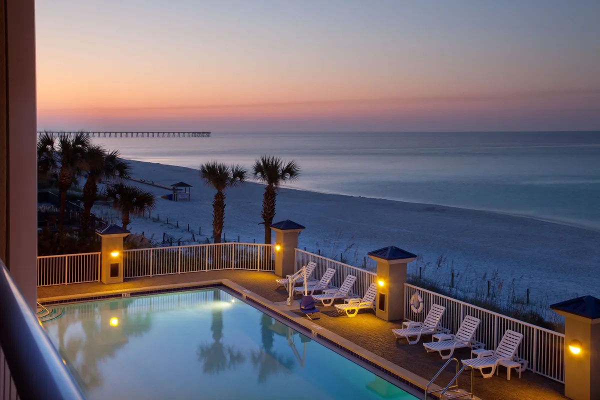 An illuminated outdoor pool sparkles under soft evening light, framed by white lounge chairs and swaying palm trees. In the background, the beach and ocean horizon glow subtly, evoking a sense of relaxation and indulgence. The scene hints at leisurely swims, poolside relaxation, and transitions from vibrant daytime to romantic evenings.