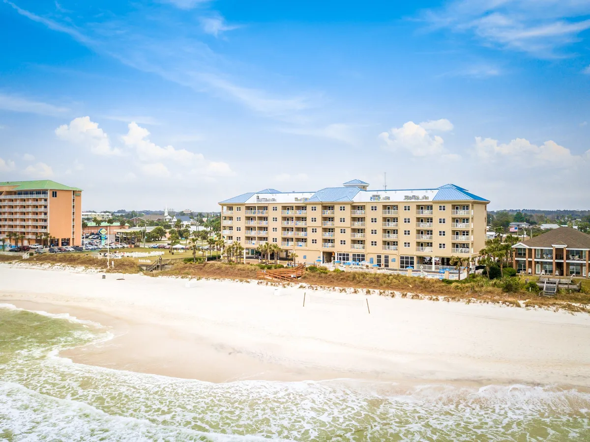 A charming beachfront villa with a blue roof stands steps from a sweeping expanse of white sand and rolling ocean waves. Palm trees and adjacent buildings suggest a lively coastal setting, while villa balconies face the Gulf, offering stunning water views. The atmosphere is refreshing and uplifting, ideal for both quiet mornings and vibrant evenings.
