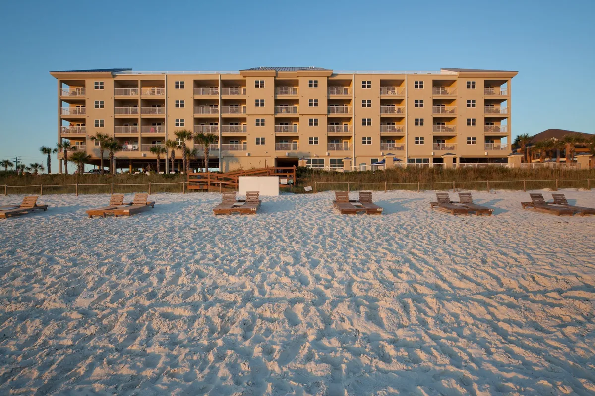A contemporary beachfront villa building stands behind rows of wooden lounge chairs arranged on soft sand, with a bright blue sky overhead. The scene showcases the villa’s immediate access to the beach and its serene, inviting ambiance.