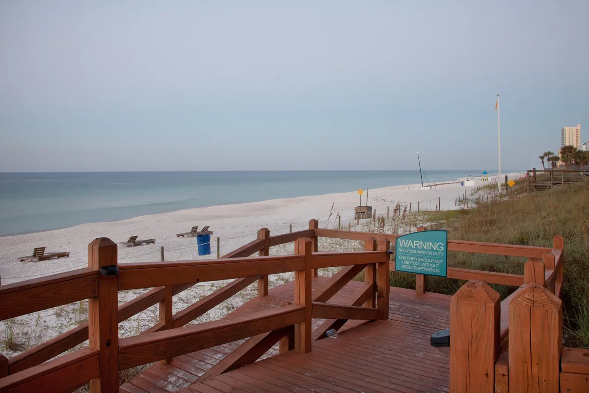 A sun-kissed wooden boardwalk leads directly through soft, powdery white sand to the pristine shoreline. The calm blue Gulf waters stretch to the horizon, with a few beach chairs spaced along the shore, suggesting both privacy and tranquility. The scene is calm and inviting, ideal for couples seeking serene mornings or romantic sunset strolls just steps from their accommodation.