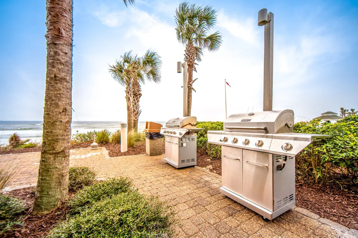 A paved patio area sits just steps from the beach, featuring two modern stainless steel BBQ grills surrounded by palm trees. The area offers sweeping ocean views, inviting guests to gather for outdoor meals and socializing in a picturesque, beachside setting.