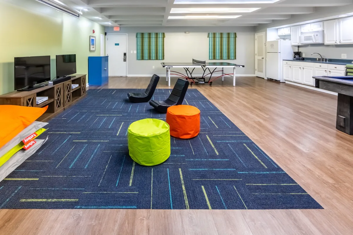 A playful, multi-use interior space with a ping pong table at the center, two black floor chairs, colorful round poufs, and a large flat-screen TV. The kitchen area is visible in the background, equipped for preparing snacks or drinks, emphasizing the space's versatility for both fun and food-related activities.