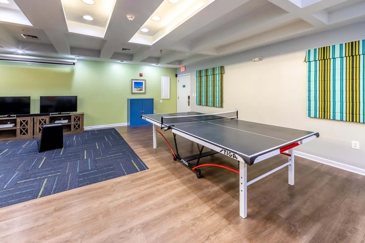 An interior games room featuring a ping pong table, two flat-screen TVs, and a blue cabinet, with striped wall decorations adding a playful touch. The space is designed for energetic activities and group entertainment, offering a lively contrast to the relaxing pool area.
