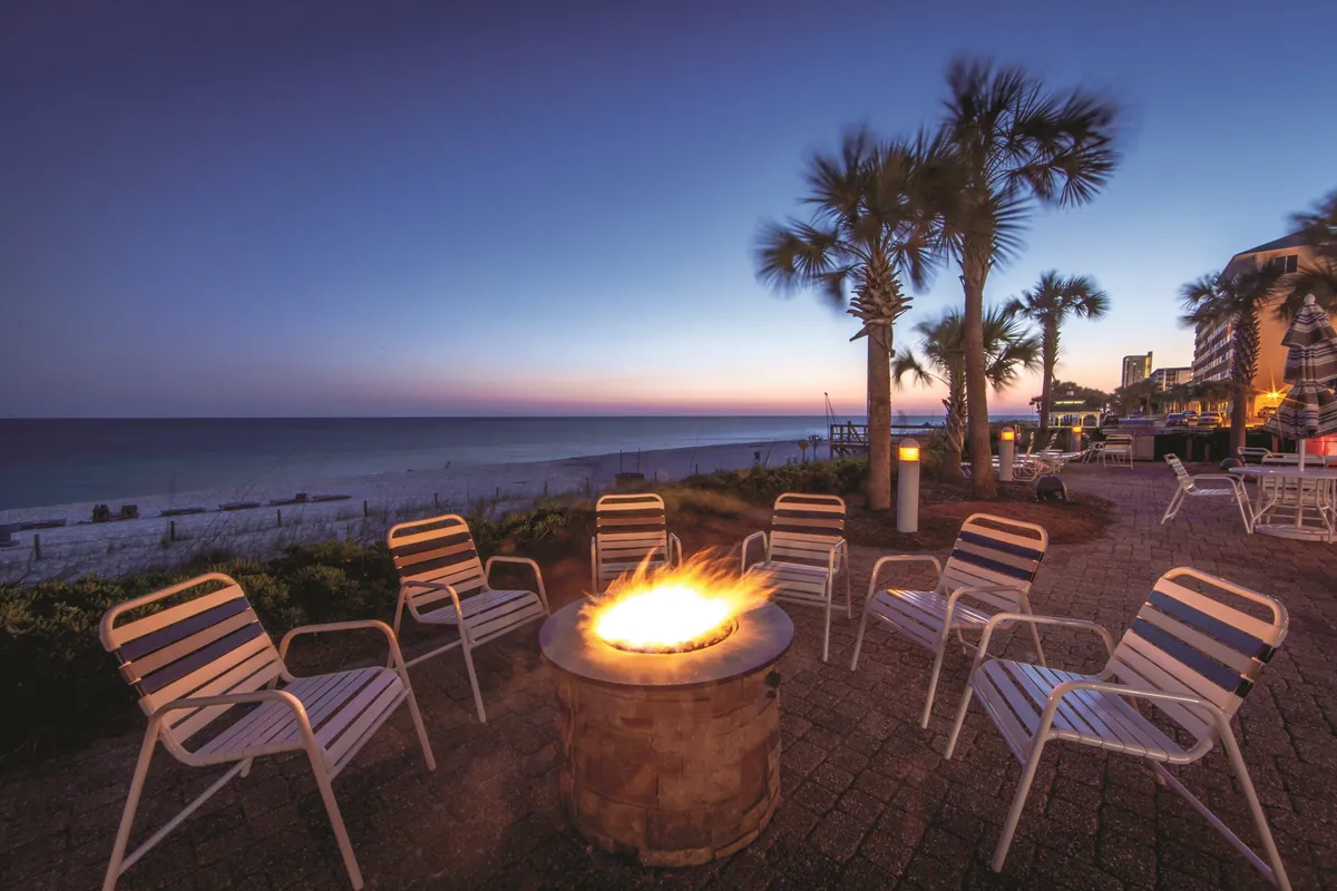 A circular outdoor fire pit surrounded by white lounge chairs, with palm trees silhouetted against a sunset sky and views of the calm ocean and sandy beach. The cozy fire pit area invites guests to gather for evening socializing, storytelling, and s’mores.
