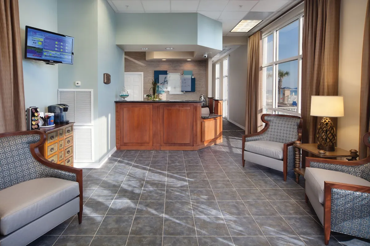 The image shows a welcoming interior space featuring a wooden reception desk, comfortable armchairs, large windows with flowing curtains, and a wall-mounted TV. The area is bathed in natural light, creating an inviting and orderly atmosphere, with a professional yet homely feel.