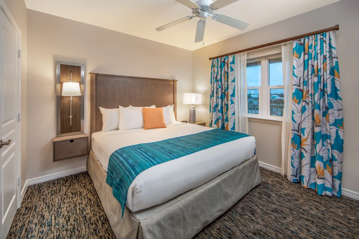 A serene bedroom with a king-sized bed dressed in a turquoise throw, floral curtains, and a nightstand with a lamp. The room is bathed in soft natural light, offering a tranquil atmosphere for restful nights.